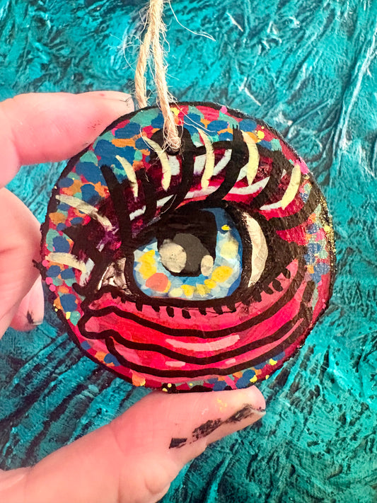 A hand holds a round, hand-painted wooden ornament shaped like a stylized eye, featuring vibrant red, pink, blue, yellow, and black painted details with a natural jute hanging string. The background is a textured turquoise blue surface.