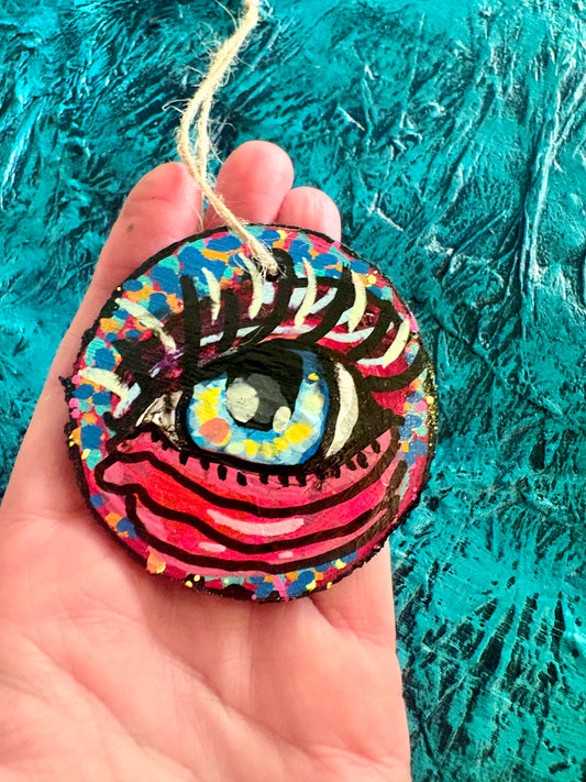 A hand holding a round, hand-painted wooden ornament featuring a stylized eye with a blue iris, black eyelashes, and pink and red stripes, set against a textured teal background.