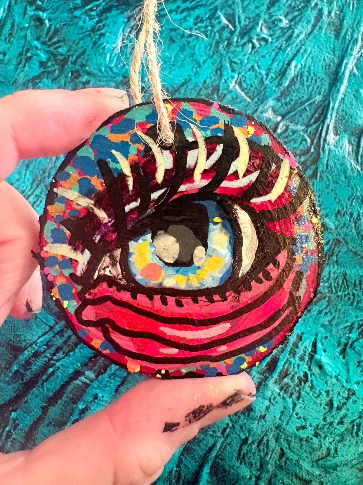 A hand holds a round, hand-painted wooden ornament shaped like a stylized eye, featuring vibrant red, pink, blue, yellow, and black painted details with a natural jute hanging string. The background is a textured turquoise blue surface.