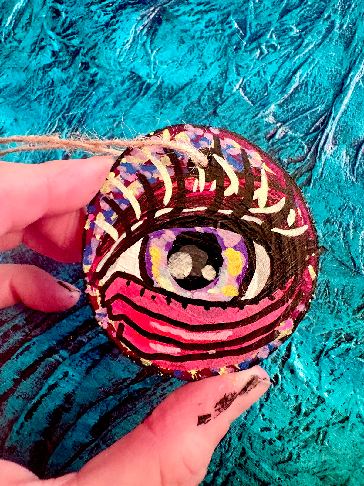 A hand holding a round, hand-painted wooden ornament featuring a stylized eye with pink, red, purple, yellow, and black details, set against a textured turquoise background.