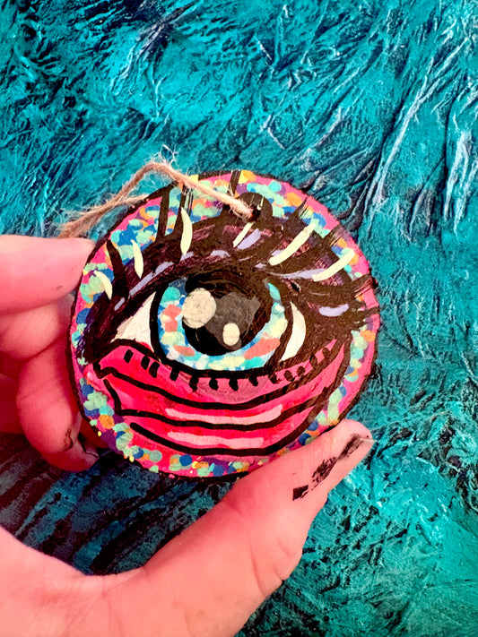 A hand holding a round, hand-painted wooden ornament featuring a stylized eye with black lashes, a pink and red mouth, and a multicolored speckled background, set against a textured teal painted backdrop.